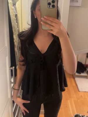 Svart ärmlös linnetopp med knyt - Snygg svart linnetopp med v-ringning och knyt framtill. Toppen är ärmlös och har en luftig, lite utsvängd passform som ger en avslappnad vibe. Perfekt att matcha med jeans eller kjol för en trendig look.