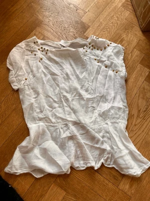 Vit blus med gulddetaljer - Snygg vit blus med korta ärmar och guldfärgade nitar vid axlarna. Blusen har en lös passform och dragkedja i ryggen samt en liten slits baktill. Perfekt till jeans eller kjol för en trendig look.Skriv gärna för mått! 💕