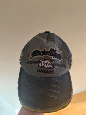 Grå keps med brodyr och slitningar - Säljer en grå keps med broderad text '1969', 'Kingston Jamaica' och 'TradeMark' framtill. Kepsen har slitna detaljer och gul kant på skärmen för en vintage look. Perfekt för dig som gillar streetwear och unik stil.