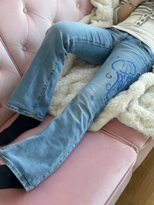 Ljusblå utsvängda jeans med manetmotiv - Unika ljusblå jeans med utsvängda ben och handritat blått manetmotiv på båda benen. Jeansen har klassisk femficksdesign och är tillverkade i denim. Perfekta för dig som vill sticka ut med ett kreativt och personligt plagg.