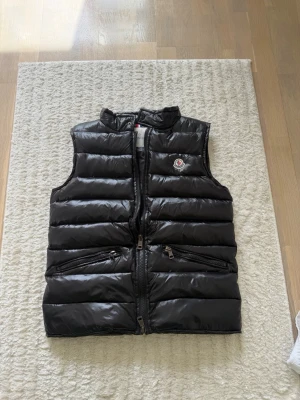 Svart dunväst från Moncler - Snygg svart dunväst från Moncler med glansig finish och klassisk logga på bröstet. Västen har två dragkedjeförsedda fickor framtill och hög krage. Perfekt för lager-på-lager och streetstyle. Stängs med dragkedja och har quiltad design. Storlek m men passar S också 