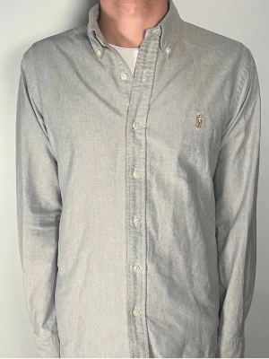 Grå Ralph Lauren skjorta - Klassisk grå skjorta från Polo Ralph Lauren i slim fit-modell. Skjortan har knappar framtill, button-down krage och den ikoniska broderade loggan på bröstet. Tillverkad i mjuk bomull och har långa ärmar. Perfekt för en clean och stilren look.