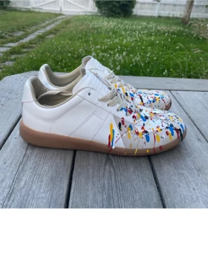 Maison Margiela sneakers med färgstänk - Unika vita sneakers från Maison Margiela med färgstänk i blått, rött, gult och svart. Skorna har beige sula och vita skosnören. Tillverkade i läder med klassisk låg siluett och cool konstnärlig design. Perfekta för dig som vill sticka ut med din stil.