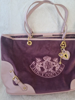 Juicy Couture scottie tote  - Snygg lila handväska från Juicy Couture i mockaliknande material med ljusrosa detaljer i skinnimitation. Väskan har guldfärgade metalldetaljer, dubbla handtag och en dekorativ nyckelberlock. Stor broderad logga framtill ger en lyxig känsla. Finns en lite defekt i hörnet ( visa på bild )