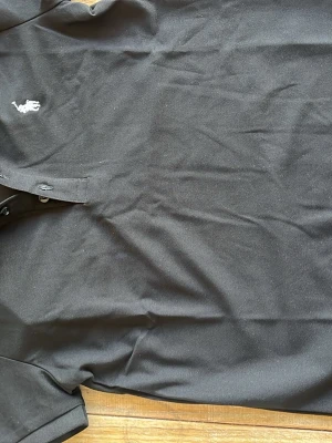 Svart pikéskjorta från Ralph Lauren - Klassisk svart pikéskjorta från Polo Ralph Lauren med vit broderad logga på bröstet. Skjortan har krage, knappar framtill och är tillverkad i mjuk bomull. Perfekt för en clean och stilren look. Storlek S men den är mindre i storlek så passar folk med XS