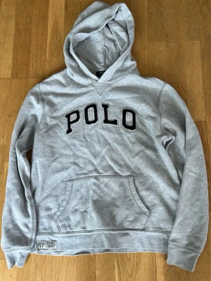 Grå hoodie från Polo Ralph Lauren - Snygg grå hoodie från Polo Ralph Lauren med stor svart POLO-text broderad på bröstet. Klassisk känguruficka framtill och huva. Tillverkad i mjuk bomullsblandning, perfekt för chill dagar eller när du vill ha en clean streetstyle-look. Passar även S. Priset är inte satt i sten!