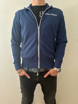 Palm Angels track jacket - Säljer en marinblå hoodie från Palm Angels med vit dragkedja och logga på bröstet. Hoodien har vita ränder längs ärmarna och ribbade muddar. Autenticitets bevis finns!