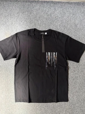 Amiri t-shirt - Snygg svart t-shirt från AMIRI med stort tryck på ryggen och mindre tryck på bröstet. Trycket består av AMIRI-loggan och färgstänk i blått, orange och vitt som rinner ner. Klassisk passform och rund hals. Perfekt för dig som vill sticka ut. Pris går diskuteras vid snabb affär