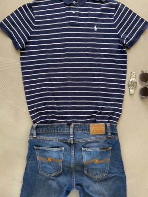 Ralph Lauren | Piké | L - Denna pikétröja för män har en klassisk och stilren design i blått med vita ränder. Den är tillverkad i ett mjukt och bekvämt material som andas bra och ger en skön pajssform hela dagen. Med sin tidlösa look passar den lika bra till vardags som vid mer uppklädda tillfällen och är enkel att matcha med både jeans och chinos.