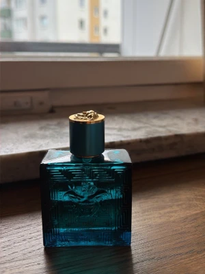 Versace Eros Eau de Parfum 100ml - Säljer en Versace Eros Eau de Parfum i en lyxig blå glasflaska med gulddetaljer och klassisk Medusa-logga på framsidan. Flaskan har en fyrkantig form och ett elegant lock med guldfärgad topp. Perfekt för dig som gillar exklusiva dofter.