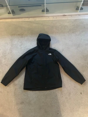 Svart vindjacka från The North Face - Snygg svart vindjacka från The North Face med huva och dragkedja framtill. Modellen heter M Quest Jacket och har en minimalistisk design med logga på bröstet. Perfekt för blåsiga dagar. Tveka inte om ni har några frågor.