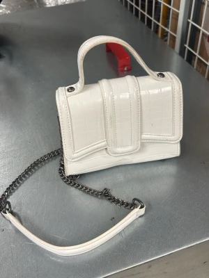Vit minihandväska med kedja - Snygg vit minihandväska i croco-mönstrat konstläder med både handtag och axelrem i silverfärgad kedja. Väskan har en unik formad lockdetalj och stilrena metalldetaljer. Perfekt accessoar för att lyfta din outfit.