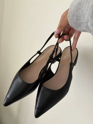 Svarta ballerinaskor med spetsig tå - Stilrena svarta ballerinaskor med spetsig tå och öppen häl. Skorna har tunna justerbara remmar runt ankeln och är tillverkade i ett slätt, matt material som liknar skinn. Perfekta för en trendig och minimalistisk look. Använda 1 gång 
