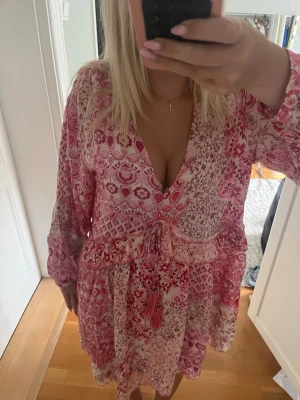 Zara klänning  - Söt kortklänning i rosa och vitt med blommigt mönster och v-ringning. Klänningen har långa ärmar, volangdetaljer och knyt i midjan med tofsar. Perfekt för sommarens varma dagar.