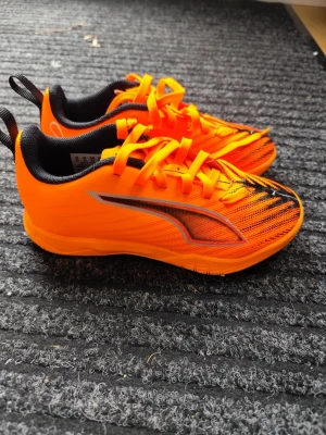 Orangea fotbollsskor från Puma - Säljer ett par riktigt snygga fotbollsskor från Puma i en skarp orange färg med svarta detaljer och snörning. Skorna har en låg profil, svart innersula och är tillverkade i syntetmaterial för bra grepp och kontroll på planen.