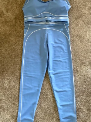 Blått träningsset från Alo - Snyggt blått träningsset från Alo med matchande sport-bh och tights. Setet har vita kontrastlinjer och logga på både bh och midja. Materialet är stretchigt och passar perfekt för yoga, gym eller annan träning.