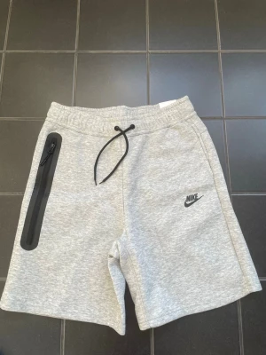Gråa Nike shorts med snörning - Snygga ljusgrå shorts från Nike med svart snörning i midjan och en praktisk dragkedjeficka på sidan. Tillverkade i mjuk bomullsmix och har Nike-logga broderad framtill. Perfekta för chill eller träning.