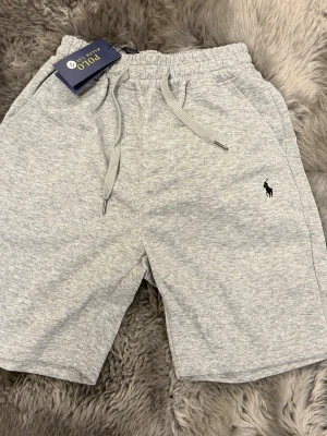 Grå shorts från Polo Ralph Lauren - Snygga ljusgrå shorts från Polo Ralph Lauren med dragsko i midjan och klassisk broderad logga på benet. Tillverkade i mjuk bomullsmix för maximal komfort. Perfekta för sommardagar eller chill hemma.