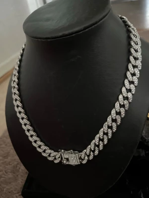 Silverfärgat cuban chain halsband - Säljer ett chunky cuban chain halsband i silverfärg med glittrande stenar på varje länk. Halsbandet har en bred design och ett lås dekorerat med små stenar för extra bling. Perfekt för dig som vill sticka ut med en statement-accessoar.