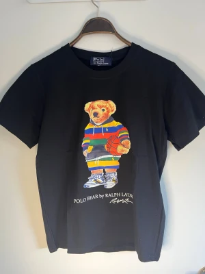 Svart Polo Bear t-shirt från Ralph Lauren - Svart t-shirt från Polo Ralph Lauren med ikoniska Polo Bear-trycket framtill. Björnen har färgglada kläder och håller en basketboll. T-shirten har rund hals och korta ärmar. Tillverkad i mjuk bomull för en skön känsla.