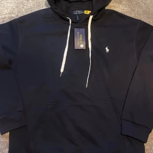 Svart hoodie från Polo Ralph Lauren - Storlek L med sitter som M Snygg svart hoodie från Polo Ralph Lauren med vit broderad logga på bröstet och vita dragsnören i huvan. Klassisk känguruficka framtill och ribbade muddar vid ärmar och nederkant. Perfekt för en chill och stilren look. (R.e.p)