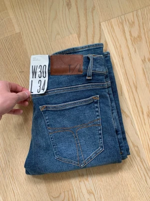 Blå tight leg jeans från Tiger of Sweden - Snygga blå slim fit jeans från Tiger of Sweden med klassisk femficksdesign och bruna sömmar. Jeansen har en läderpatch bak i midjan och är tillverkade i stretchigt denim för en bekväm passform. Perfekta för en trendig och avslappnad look.