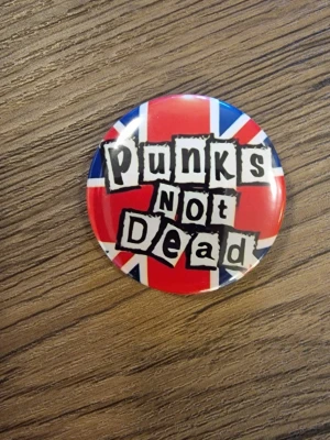 Punks Not Dead pinsknapp - Cool knapp med texten 'Punks Not Dead' i svart graffiti-stil. 4,4 cm. Perfekt accessoar för dig som gillar punk och vill sticka ut. Rund form och klassisk punkkänsla.
