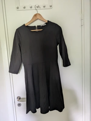 Svart klänning från Gina Tricot - En enkel och stilren svart klänning från Gina Tricot med trekvartsärm och rund halsringning. Klänningen har en lätt utställd kjol och dragkedja i ryggen. Perfekt basplagg som är lätt att styla.