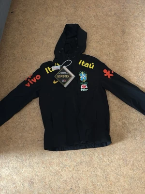 Svart Nike Brasil vindjacka Gore-Tex - Snygg svart vindjacka från Nike med Brasilien CBF-logga och sponsorer i gult och rött. Jackan har huva, hel dragkedja och är tillverkad i slitstarkt Gore-Tex-material. Perfekt för dig som gillar sportig stil och vill visa stöd för Brasilien.