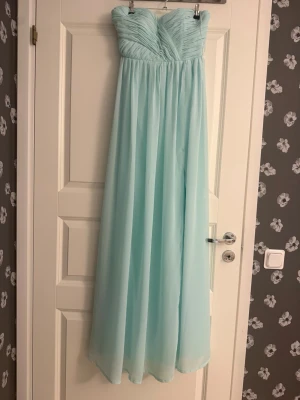 Ljusblå axelbandslös långklänning - Snygg ljusblå axelbandslös långklänning med draperad överdel och flytande kjol i tunt, svepande tyg. Perfekt för bal eller student. Klänningen har en elegant och enkel design som ger en romantisk känsla.
