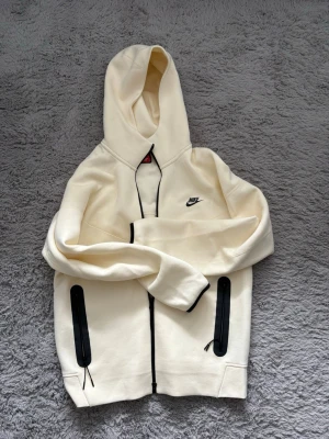 Beige hoodie från Nike med dragkedja - Snygg beige hoodie från Nike med svart logga på bröstet och hel dragkedja framtill. Två stora fickor med svarta dragkedjor och svarta detaljer längs kanterna. Tillverkad i mjukt material och har en stor huva för extra komfort.