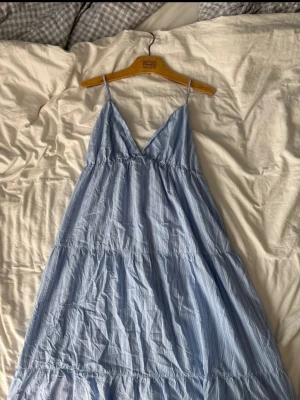 Ljusblå randig sommarklänning - Luftig långklänning från H&M i ljusblått med smala axelband och v-ringning. Klänningen har ett randigt mönster och flera volanger. Materialet känns som bomull.