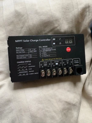 MPPT Solar Charge Controller - MPPT Solar Charge Controller i svart plast med digital display och flera anslutningar för solpanel, batteri och last. Har tydliga indikatorer för laddningsstatus och olika lägen. Passar för 12V/24V system och har inställningsknapp samt terminalblock för enkel installation.