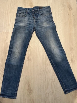 Slim fit jeans G-Star RAW - Säljer ett par blå slim fit jeans från G-Star RAW med snyggt slitna detaljer och knäppning fram. Jeansen har klassisk femficksmodell och är tillverkade i bomull med lite stretch för extra komfort. Sparsamt använda. Är 183 cm lång, har S/M. 