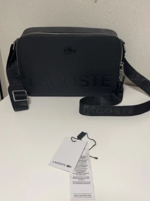 Svart Lacoste väska - Snygg svart axelväska från Lacoste med justerbar rem och präglad logotyp framtill. Väskan har en rektangulär form, dragkedja upptill och är tillverkad i slitstarkt syntetmaterial. Perfekt för att bära dina viktigaste grejer med stil.