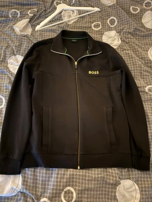 Svart zip-hoodie från BOSS - Svart hoodie från BOSS med dragkedja och två fickor framtill. Snygg vit detalj längs kragen och diskret BOSS-logga på bröstet. Perfekt för en stilren och avslappnad look. Materialet känns mjukt och bekvämt mot huden.