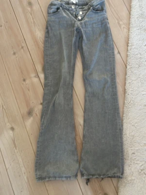 Gråa bootcut jeans från Gina Tricot - Säljer ett par gråa bootcut jeans från Gina Tricot med låg midja och flera silverfärgade knappar i gylfen. Jeansen har en avslappnad passform och råa kanter nertill för en trendig look. Perfekta för dig som gillar en retrostil.