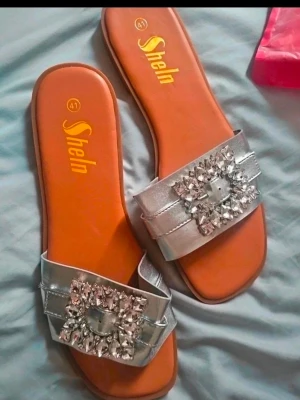 Silvriga sandaler med strass från SheIn - Glittriga sandaler från SheIn i storlek 41. De har breda, silvriga remmar i syntetmaterial och är dekorerade med stora, gnistrande strasstenar framtill. Sulan är brun och platt, vilket ger en clean och trendig look. Perfekta för att lyfta vilken outfit som helst.
