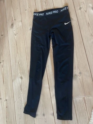 Svarta Nike Pro Dri-FIT leggings - Säljer ett par svarta Nike Pro Dri-FIT leggings med hög midja och elastisk linning där det står 'NIKE PRO'. Tillverkade i ett stretchigt funktionsmaterial som andas. Perfekta för träning eller chill. Swoosh-logga på benet för extra stil.