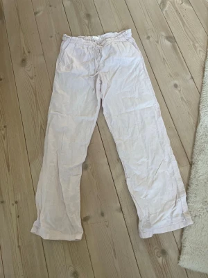 Ljusrosa linnebyxor Gina Tricot - Säljer ett par ljusrosa linnebyxor från Gina Tricot med avslappnad passform och resår i midjan. Byxorna har raka ben och är tillverkade i ett tunt, luftigt linnematerial. Perfekta för en chill och bekväm stil.
