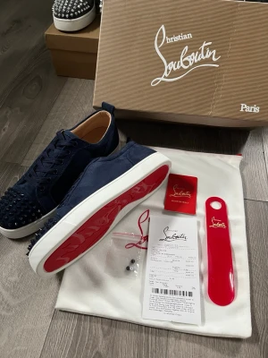 Mörkblå Christian Louboutin sneakers - Snygga mörkblå sneakers från Christian Louboutin i mocka med ikoniska röda sulor och svarta nitar på tån. Skorna har lågt skaft, vita kanter på sulan och klassisk snörning. Perfekta för dig som vill sticka ut med exklusiv design och edgy detaljer.