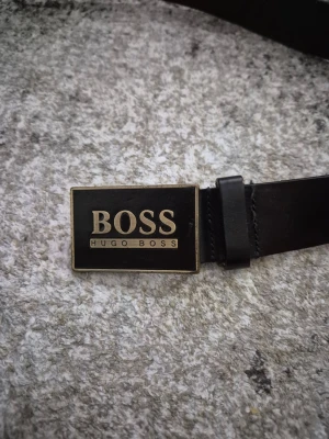 Svart skärp från Hugo Boss - Snyggt svart skärp från Hugo Boss med en rektangulär metallspänne där loggan står tydligt. Bältet är gjort i skinn och har en stilren design som passar till många olika outfits.