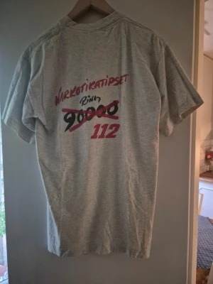 T-shirt med tryck 'Narkotikatipset' Herr M ljusgrå - Ljusgrå t-shirt med tryck fram och bak där det står 'Narkotikatipset, ring 112'. T-shirten har rund hals och korta ärmar. Perfekt för dig som gillar unika vintage-plagg. Herr stl.M
