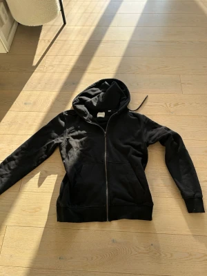 Svart zip hoodie från Jack & Jones med huva - En klassisk svart hoodie med dragkedja framtill och två stora fickor. Hoodien har en rymlig huva och ribbade muddar vid ärmar och nederkant. Perfekt för en avslappnad stil och enkel att matcha med det mesta. Materialet känns mjukt och bekvämt.
