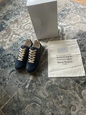 Maison Margiela blå sneakers storlek 42 - Snygga blå sneakers från Maison Margiela med vita skosnören och beige gummisula. Ovandelen är i läder och mocka, och insidan är fodrad i vitt. Klassisk låg modell med stilren design och diskreta detaljer. Perfekta för dig som gillar exklusiva och trendiga skor. Skorna är i storlek 42, helt nya och aldrig burna. Till skorna kommer en box och en dustbag. Boxen har lite små skador men som tur är inga hål.