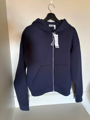 Mörkblå hoodie med dragkedja Weekday - Snygg mörkblå hoodie från Weekday med dragkedja framtill och stor känguruficka. Klassisk modell med huva och ribbade muddar vid ärmslut och nederkant. Perfekt för en avslappnad och stilren look. Tillverkad i mjuk bomullsblandning.