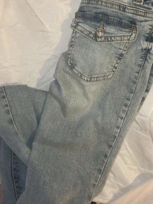 Ljusblå bootcut jeans med fickdetalj - Säljer ett par ljusblå bootcut jeans med låg midja och snygga fickdetaljer bak, inklusive lock med knapp. Jeansen har klassisk femficksdesign och är tillverkade i denim med lätt tvättad look. Perfekta för en avslappnad och trendig stil.
