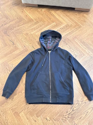 Svart hoodie från Burberry med dragkedja - Snygg svart hoodie från Burberry med dragkedja framtill och två stora fickor. Huvan har ett klassiskt rutigt foder i grått och vinrött. Tillverkad i mjukt bomullsmaterial och har justerbara snören i huvan. Perfekt för en avslappnad och stilren look.