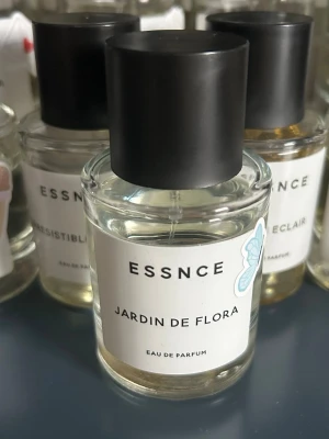 ESSNCE Jardin de Flora parfym - ESSNCE Jardin de Flora Eau de Parfum i en stilren, genomskinlig glasflaska med svart cylinderformad kork. Doften har en elegant och modern design med vit etikett och diskret blå fjärilsdetalj. Perfekt för dig som gillar fräscha och blommiga parfymer.