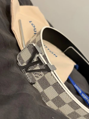 L-V rutigt bälte Damier - Säljer ett L-V bälte i Damier-mönster med klassiskt L-V-spänne i silverfärg. Bältet har rutigt mönster i vitt och grått samt svart insida. Tillverkat i skinn och detaljer i metall. Perfekt accessoar för att lyfta din outfit.